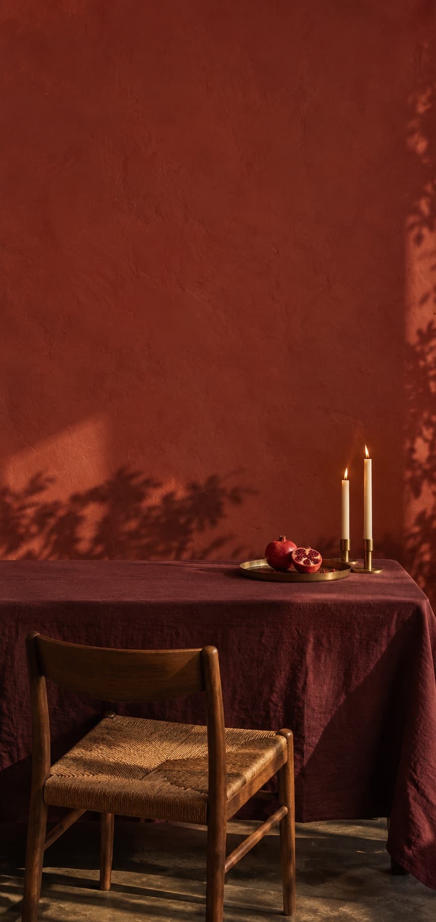A table under a wine-red cloth against a red plaster wall, with a split pomegranate, two lit candles and one empty chair.
