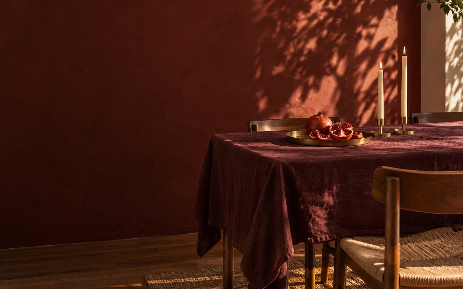 A table under a wine-red cloth against a red plaster wall, with a split pomegranate, two lit candles and one empty chair.
